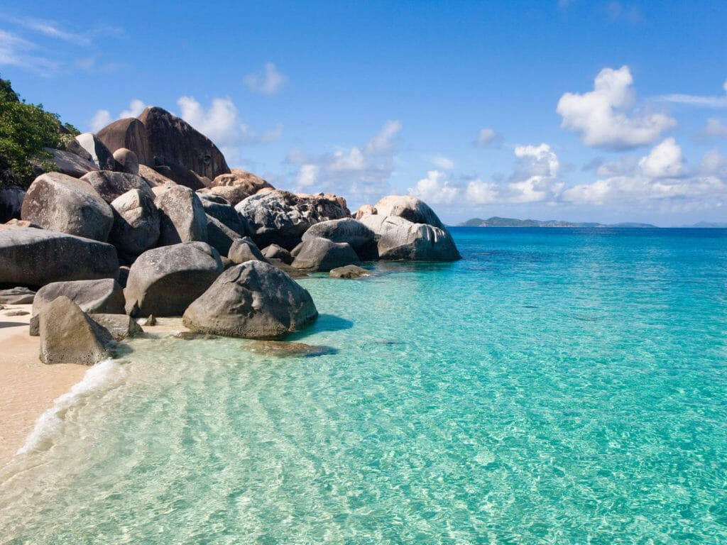 A serene view of a beach on the British Virgin Islands, captured from a distance. The image showcases the crystal-clear blue water, which gently laps against the sandy shore. The beach is framed by large rocks, which add to the natural beauty of the scene.